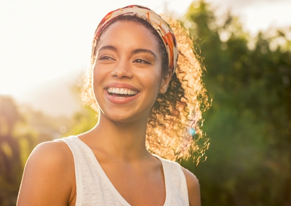 Woman smiling outdoors in natural sunlight wearing white tank top and headband.
