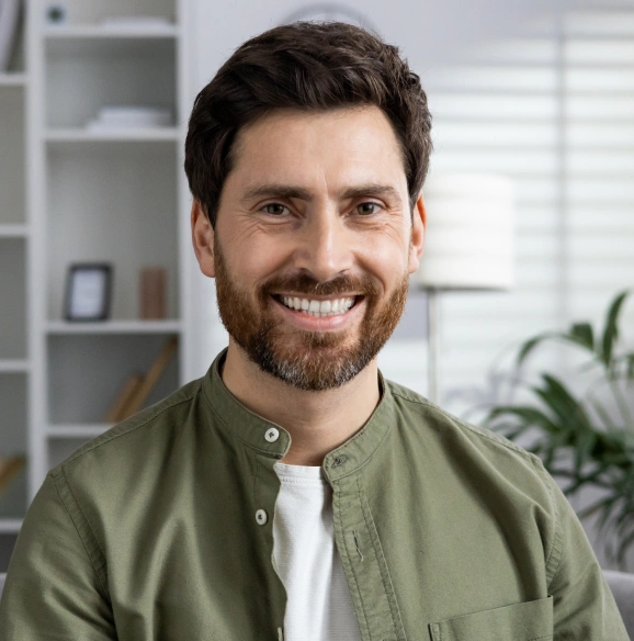Smiling man in olive green shirt in modern dental office with shelving and plants behind him.