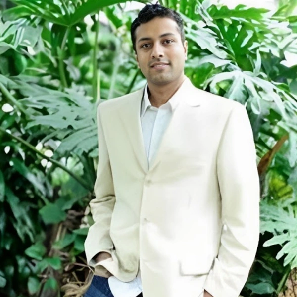 Professional man in cream blazer standing before lush green foliage backdrop.